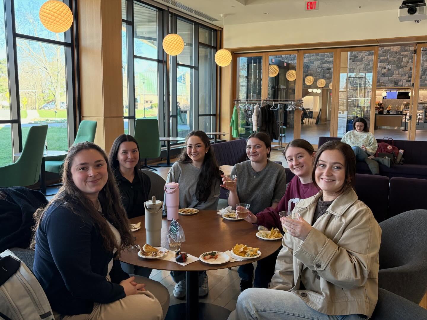 Six people seated around a table with food and drinks in a bright room with large windows and modern furniture.