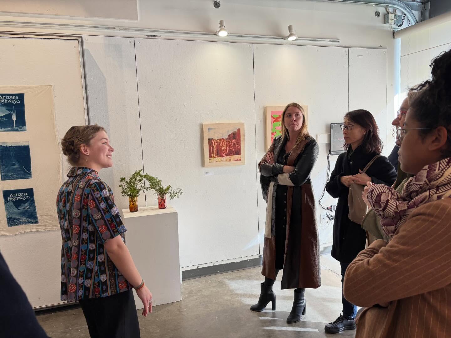 Person smiling and speaking to a group in an art gallery with paintings on the wall.
