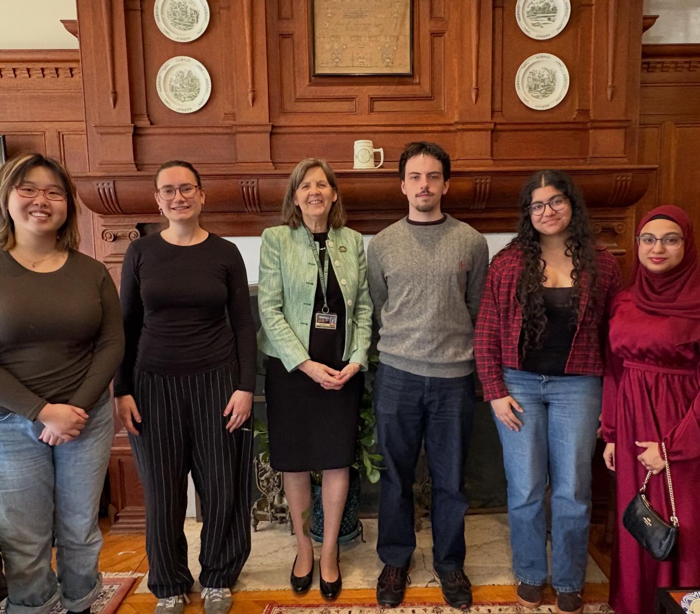 Six people standing side by side in front of a wooden fireplace with decorative plates and framed artwork above.