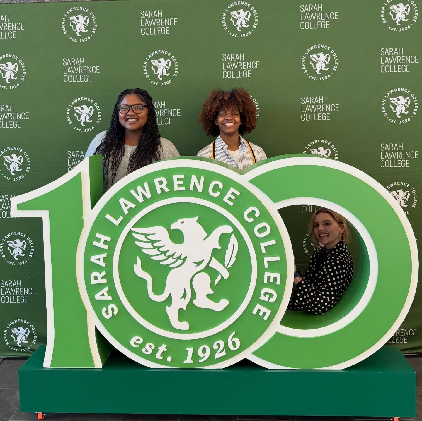 Three people pose with a large "100" sign at a Sarah Lawrence College backdrop, featuring the college's logo and establishment date, 1926.