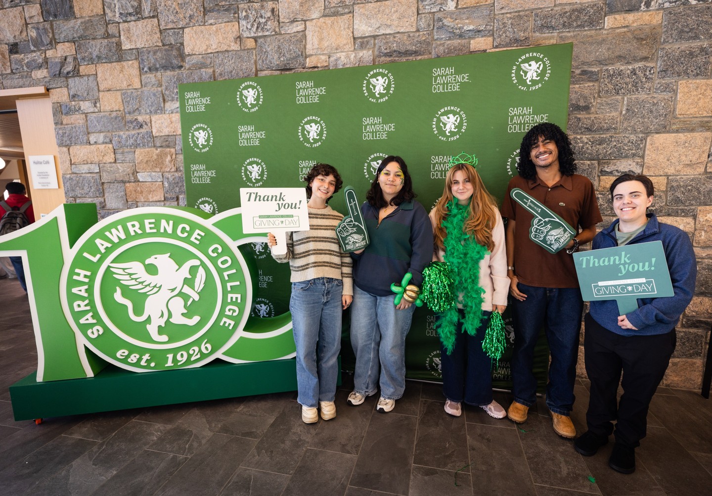 Six people standing in front of a Sarah Lawrence College backdrop, holding "Thank You" signs and green foam fingers for Giving Day.