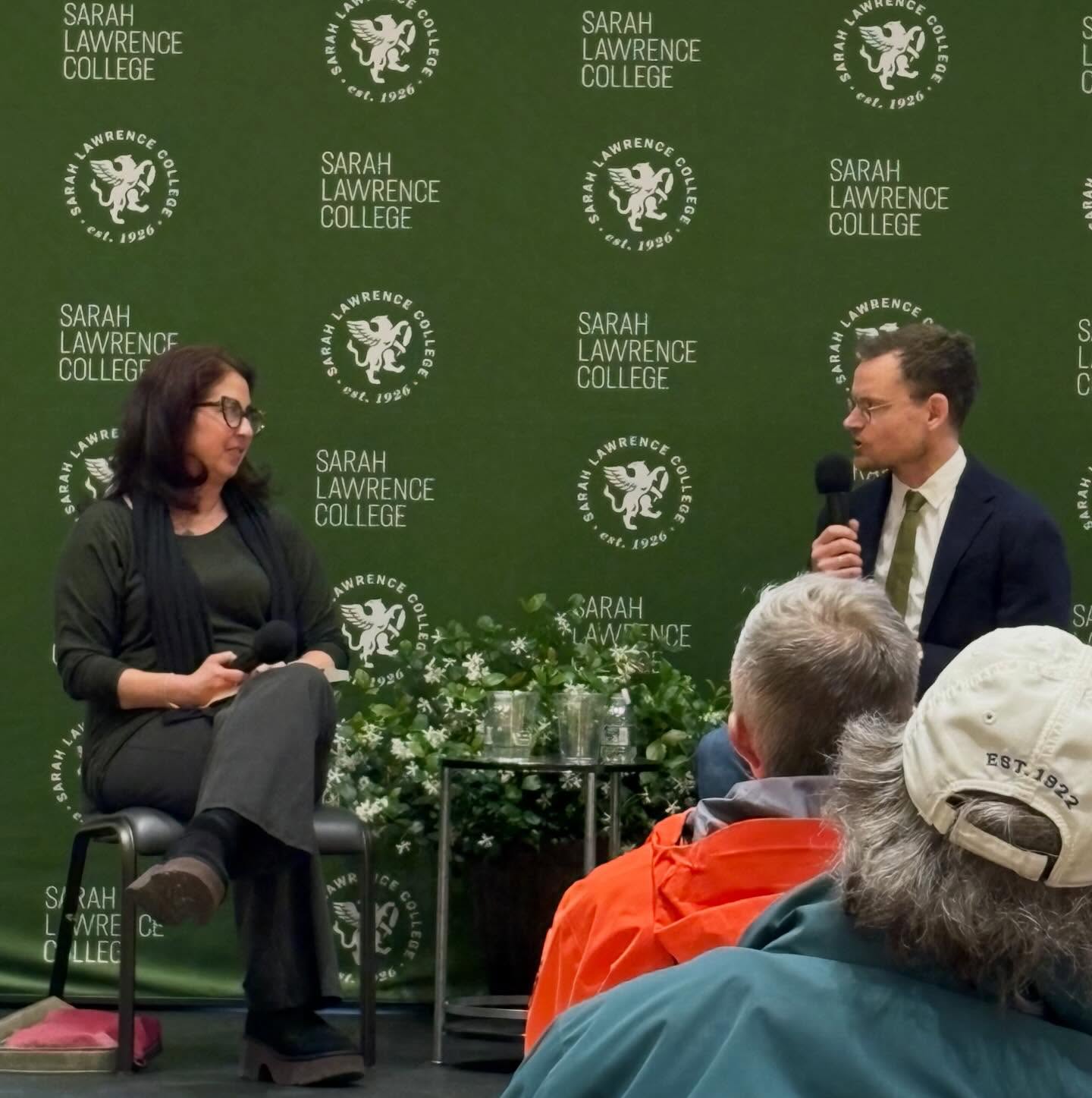 Two people seated on stage with microphones at Sarah Lawrence College event, a green backdrop and audience in foreground.