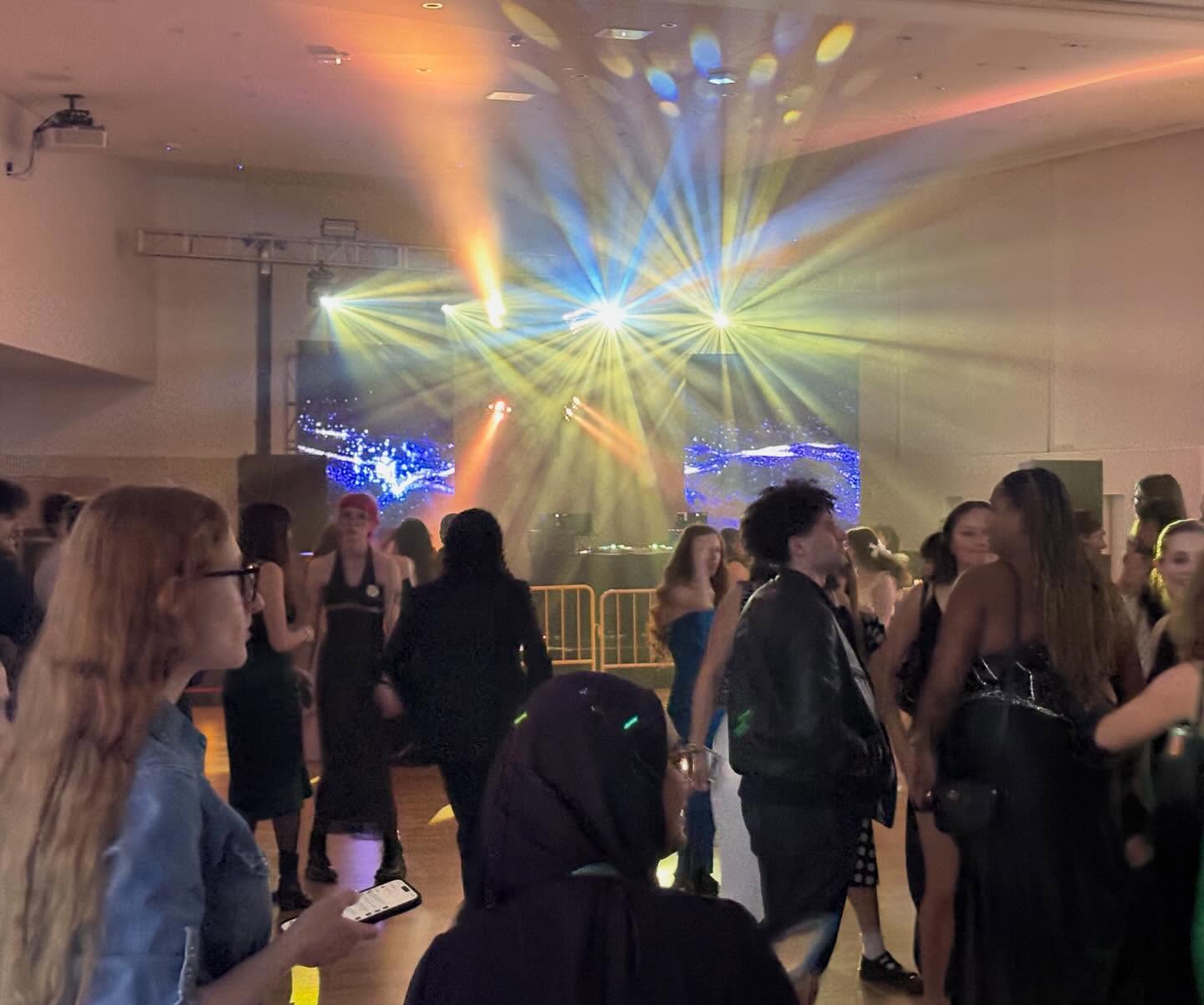 People dancing in a dimly lit room with colorful lights shining from a stage. A vibrant atmosphere with projected visuals in the background.