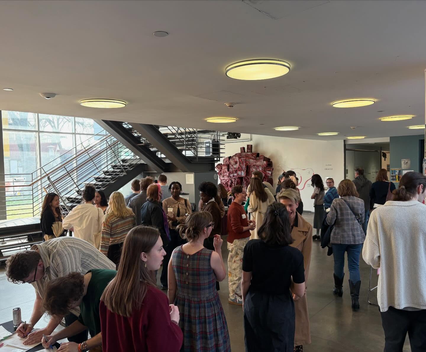 A group of people mingling and conversing in a brightly lit room with large windows and a staircase.