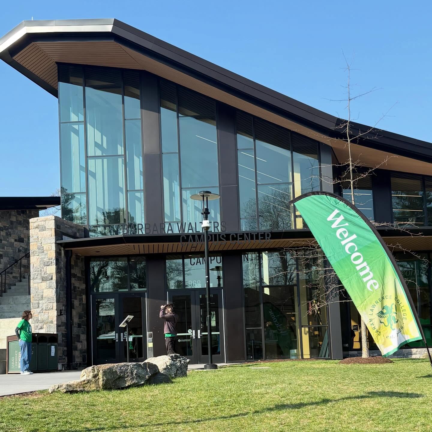 Modern campus center with large glass windows and stone walls. A green "Welcome" flag stands on the lawn near the entrance.
