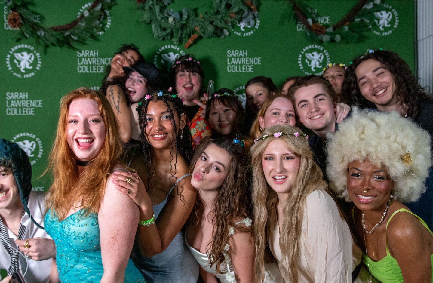 Group of smiling individuals wearing colorful outfits and flower crowns, posing in front of a green Sarah Lawrence College backdrop.