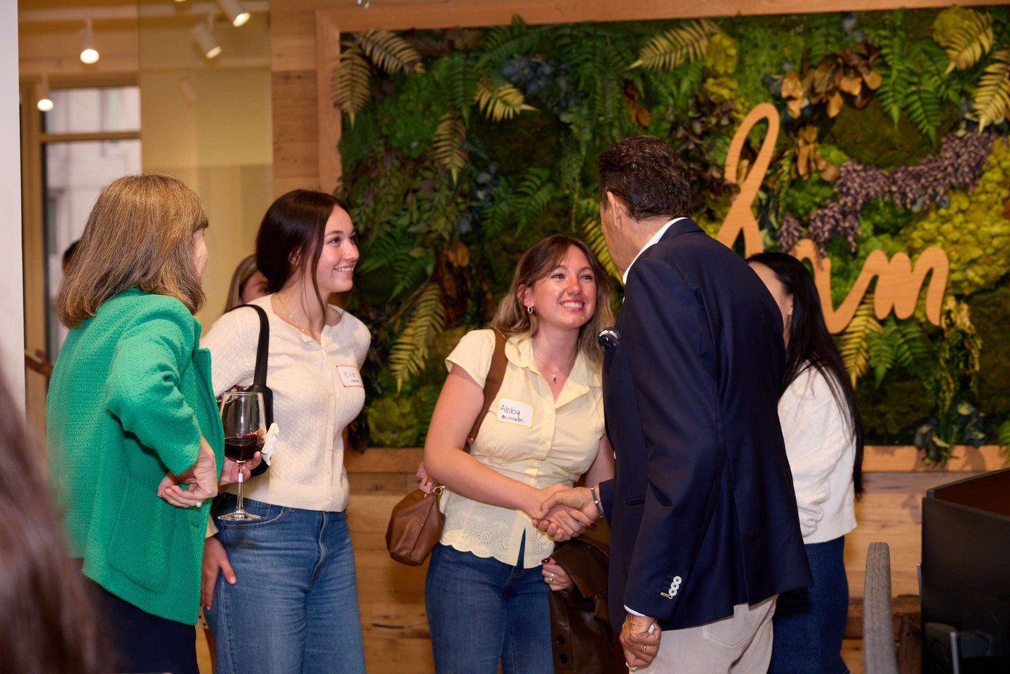 People conversing at an indoor event with a green wall backdrop. One person shakes hands while another holds a wine glass.