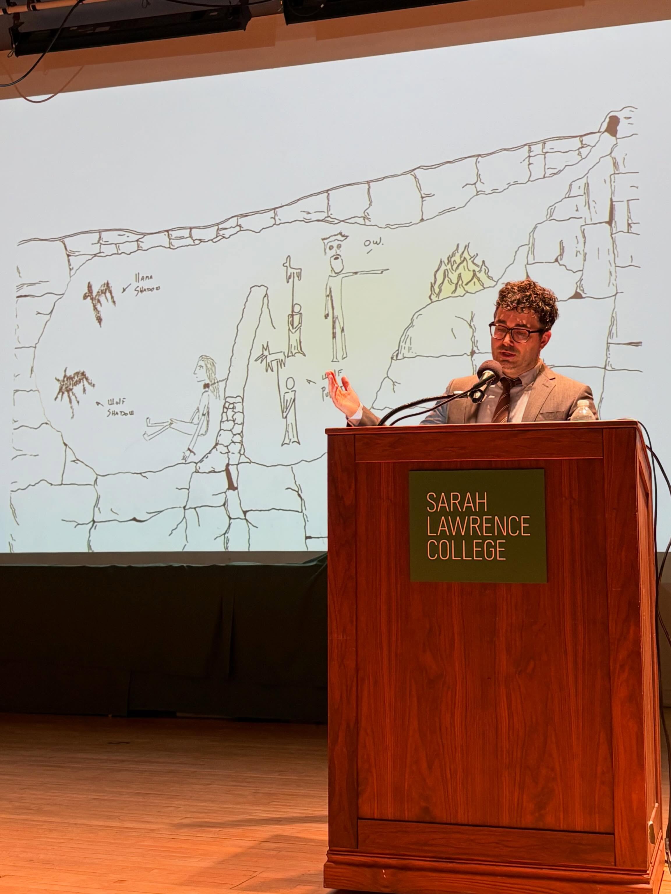Speaker at a podium with Sarah Lawrence College sign, in front of a screen displaying a drawing resembling Plato's cave allegory.
