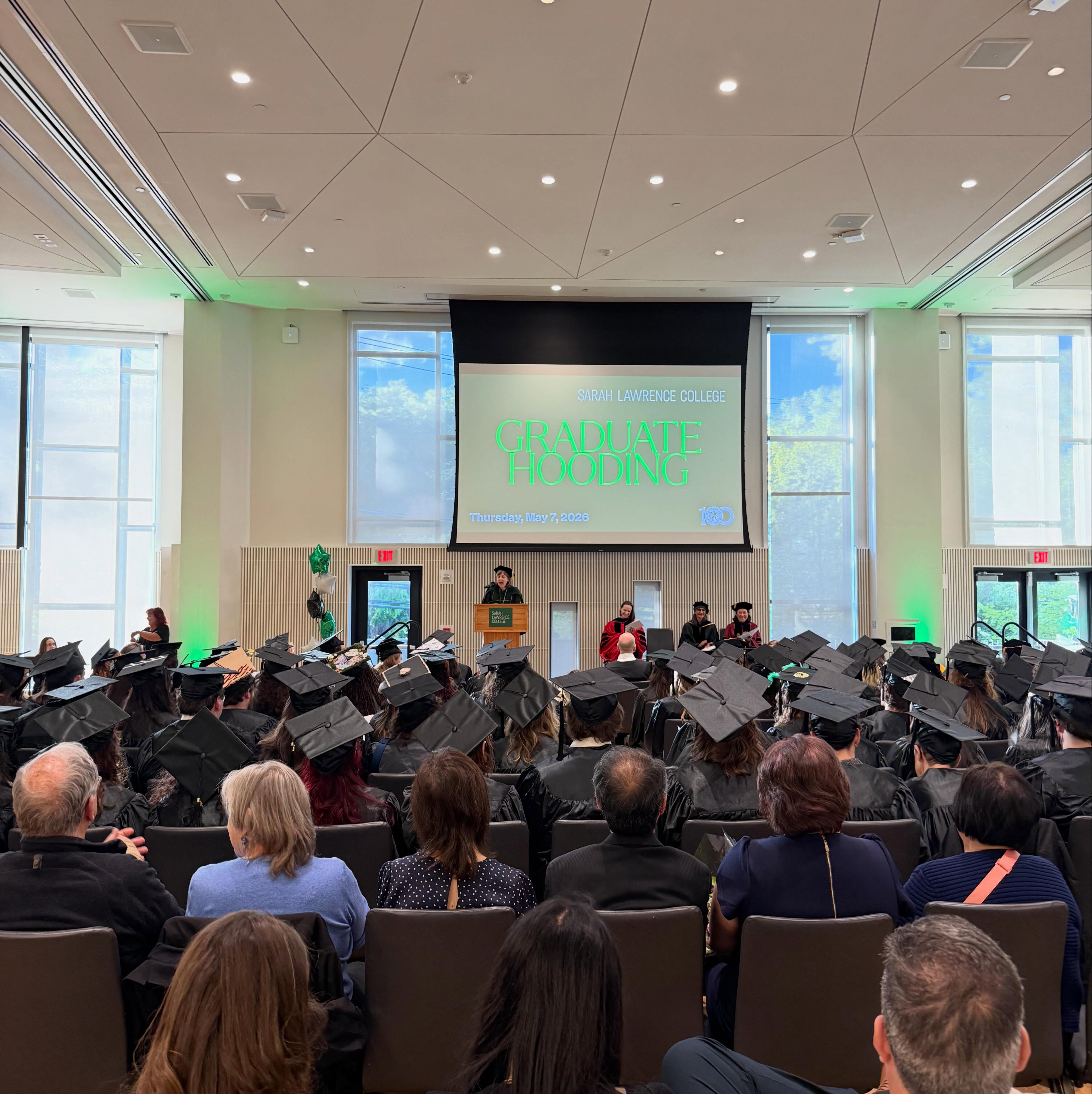 Indoor graduate hooding ceremony at Sarah Lawrence College with a speaker at the podium. Graduates in caps and gowns are seated, facing the stage.