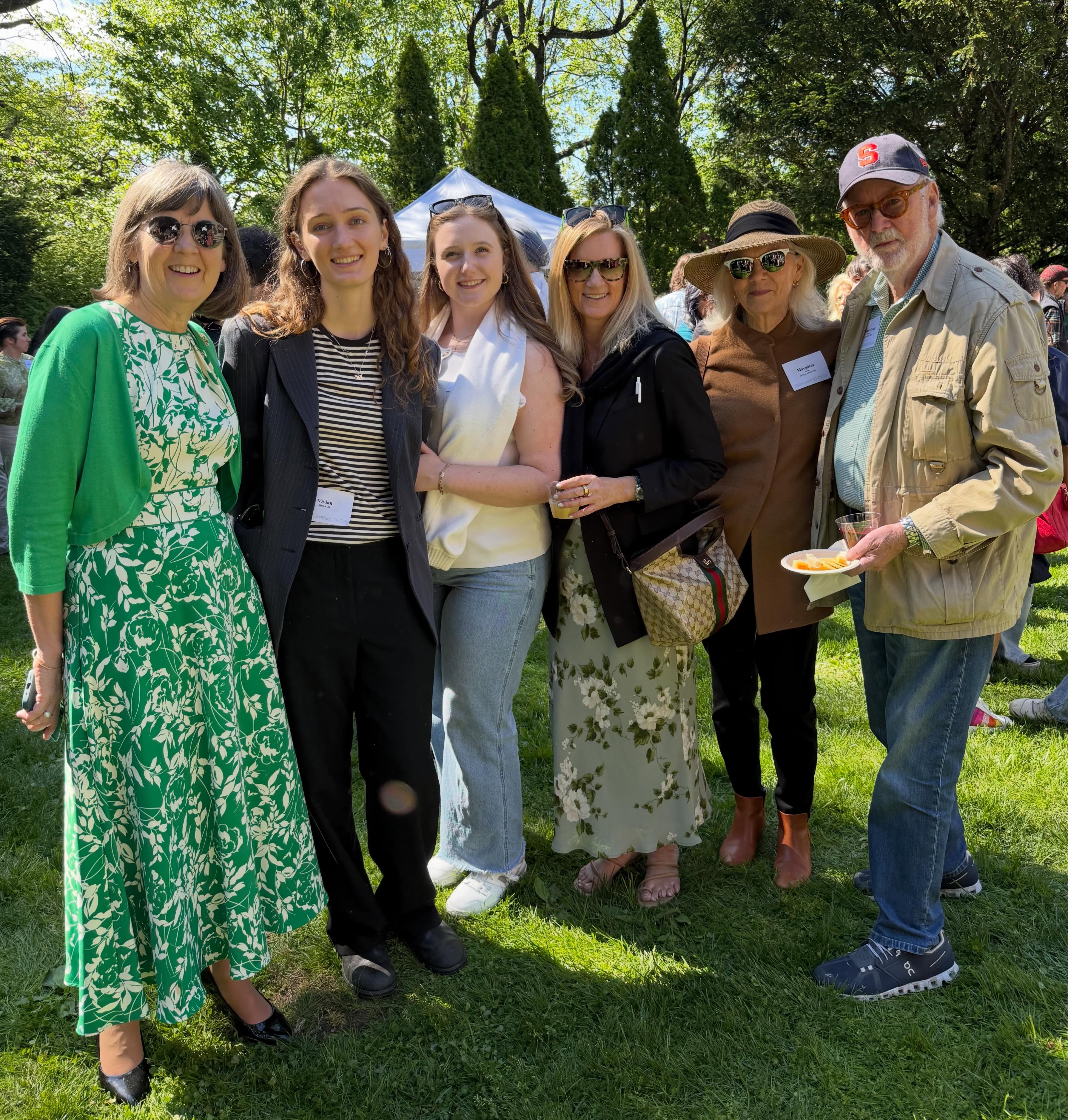 Six people smiling and standing together on grass at a sunny outdoor gathering, with trees and a tent in the background.