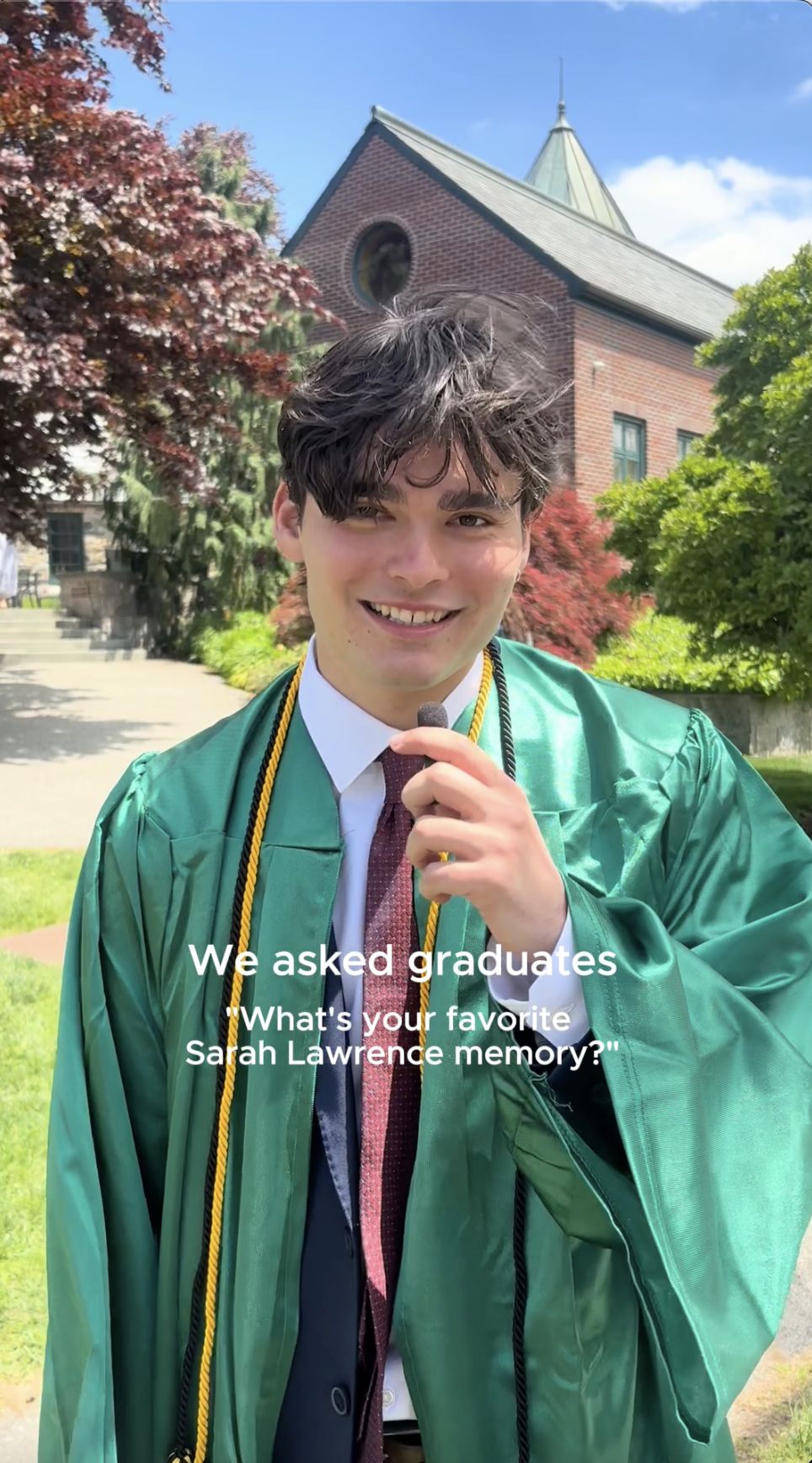 Graduate in green gown holding a microphone, with text overlay asking for a favorite Sarah Lawrence memory. A brick building is in the background.
