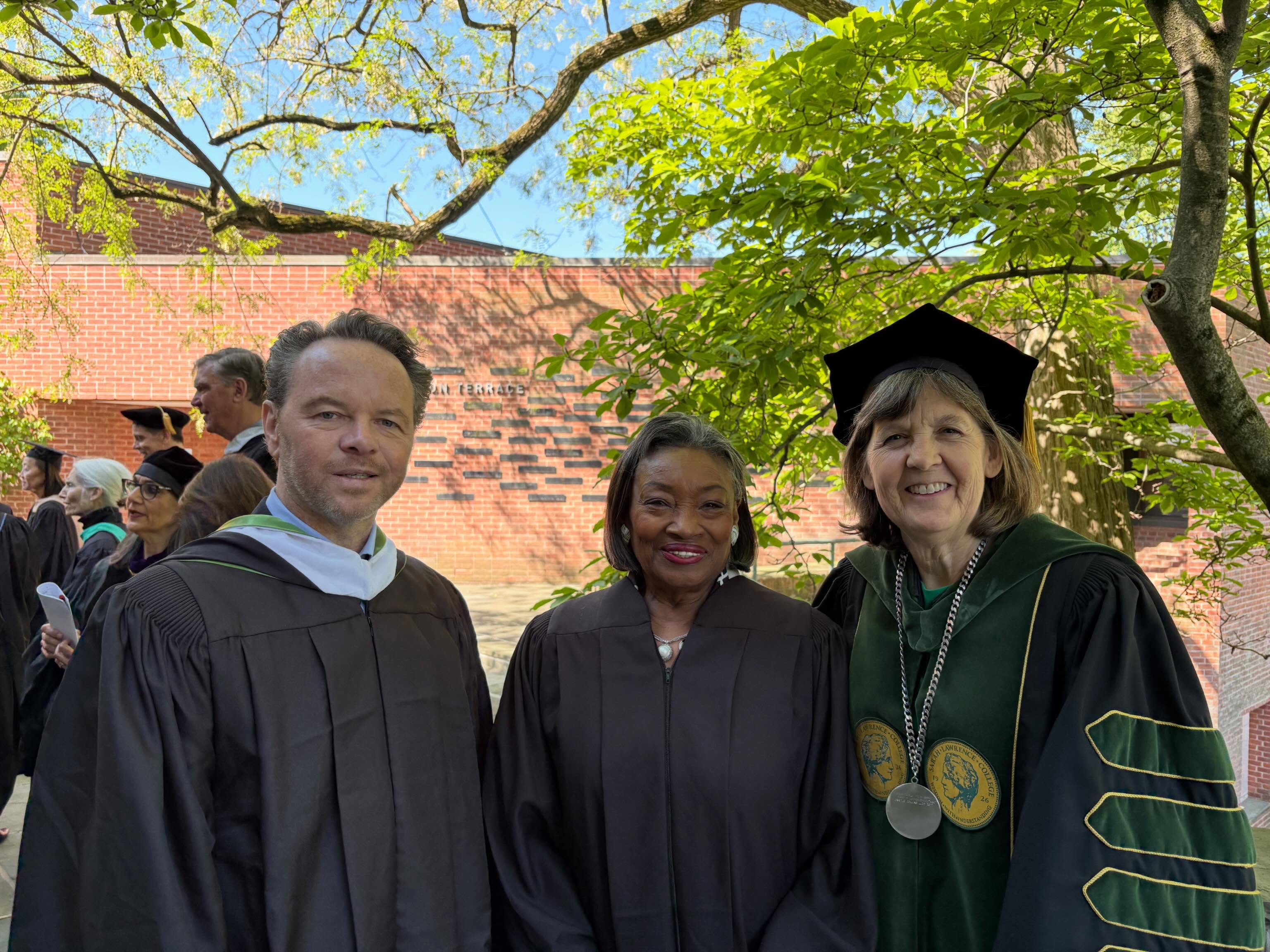 Three individuals in graduation attire stand outside, smiling. A brick wall and a leafy tree are in the background.