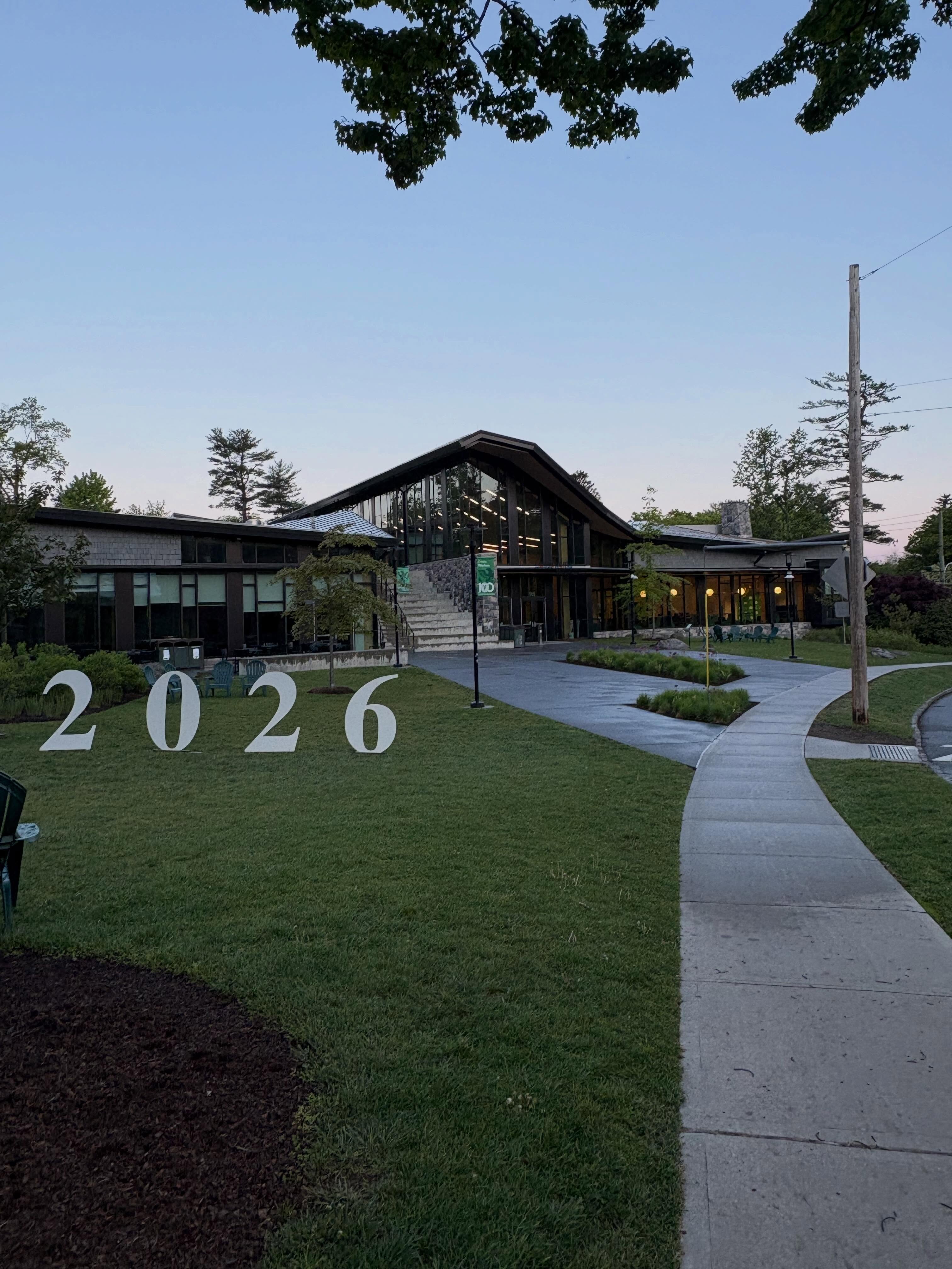 A modern building at dusk with a curved walkway and large "2026" numbers on the lawn, surrounded by trees.