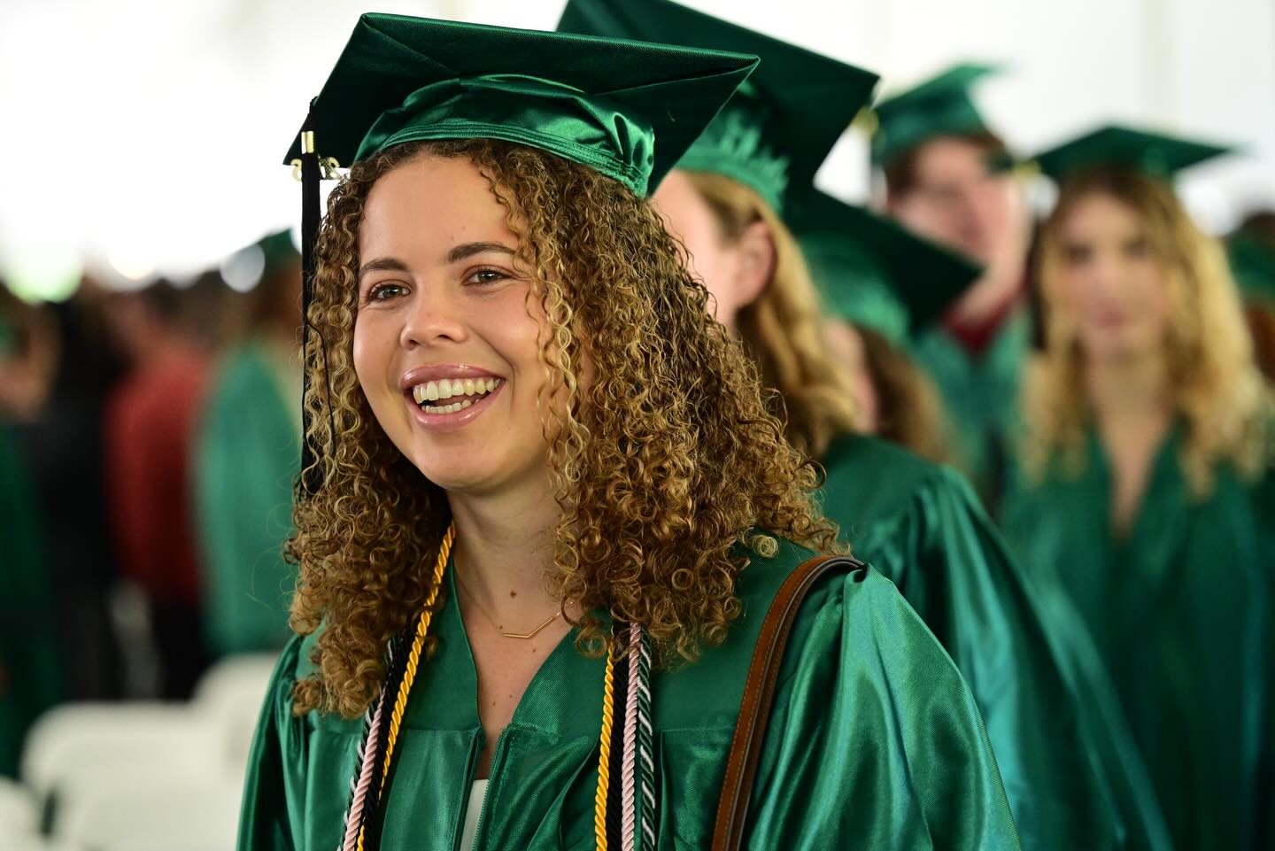 Graduate in a green cap and gown smiling at a commencement ceremony with others in the background.