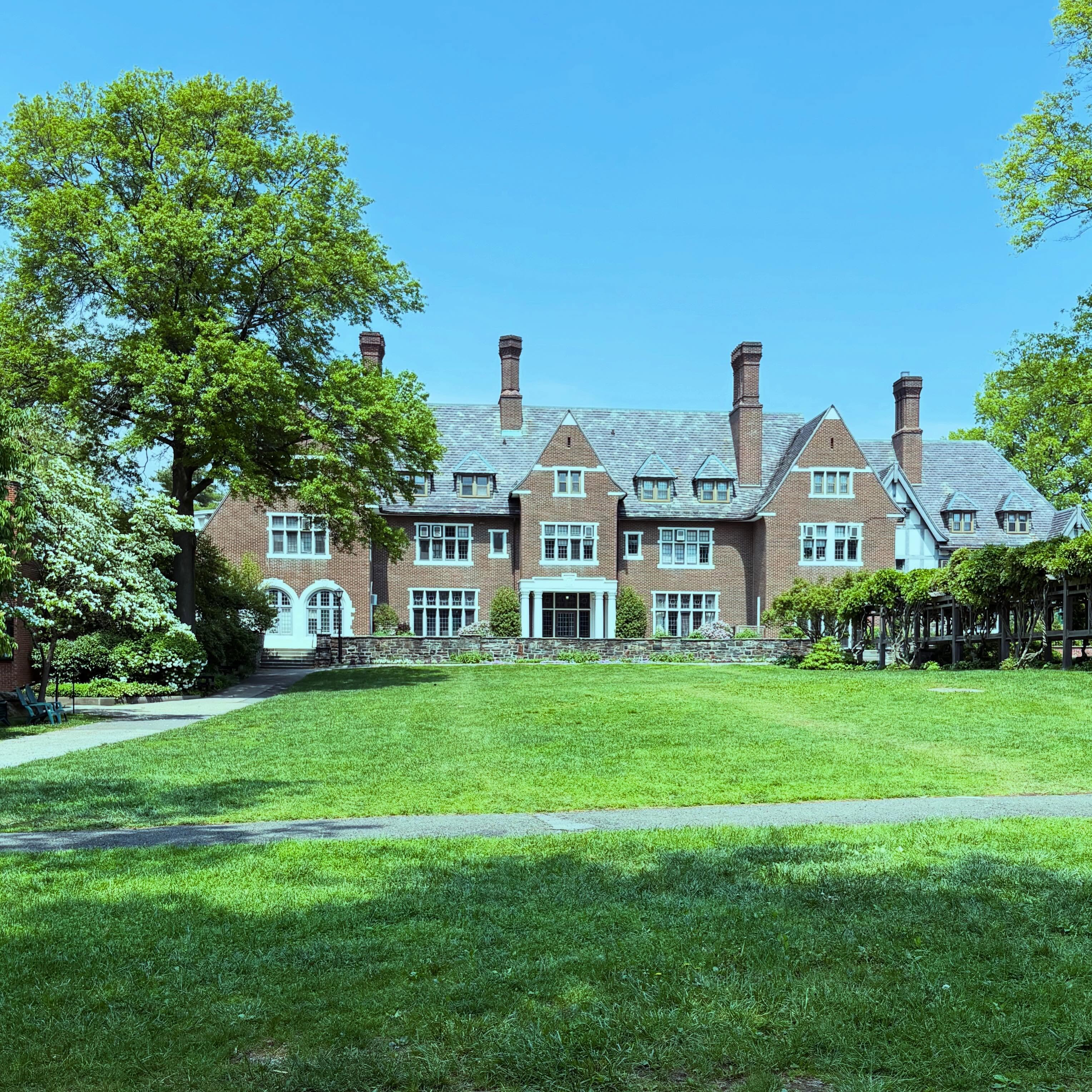 Large brick building with multiple chimneys and peaked roofs, surrounded by lush trees and a well-kept lawn under a clear blue sky.