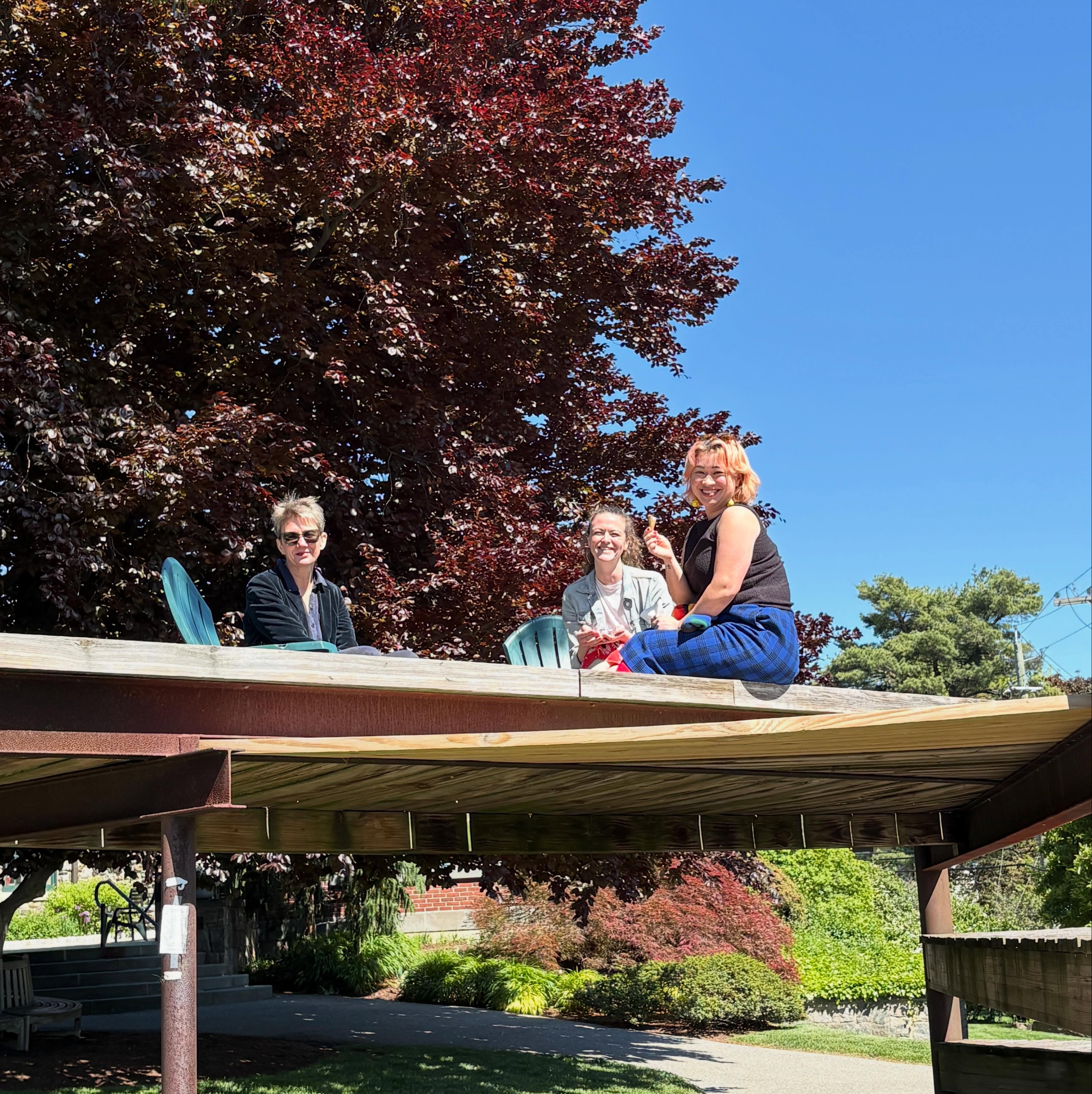 Three people sitting on a raised wooden platform outdoors, under bright blue sky, surrounded by lush trees and greenery.