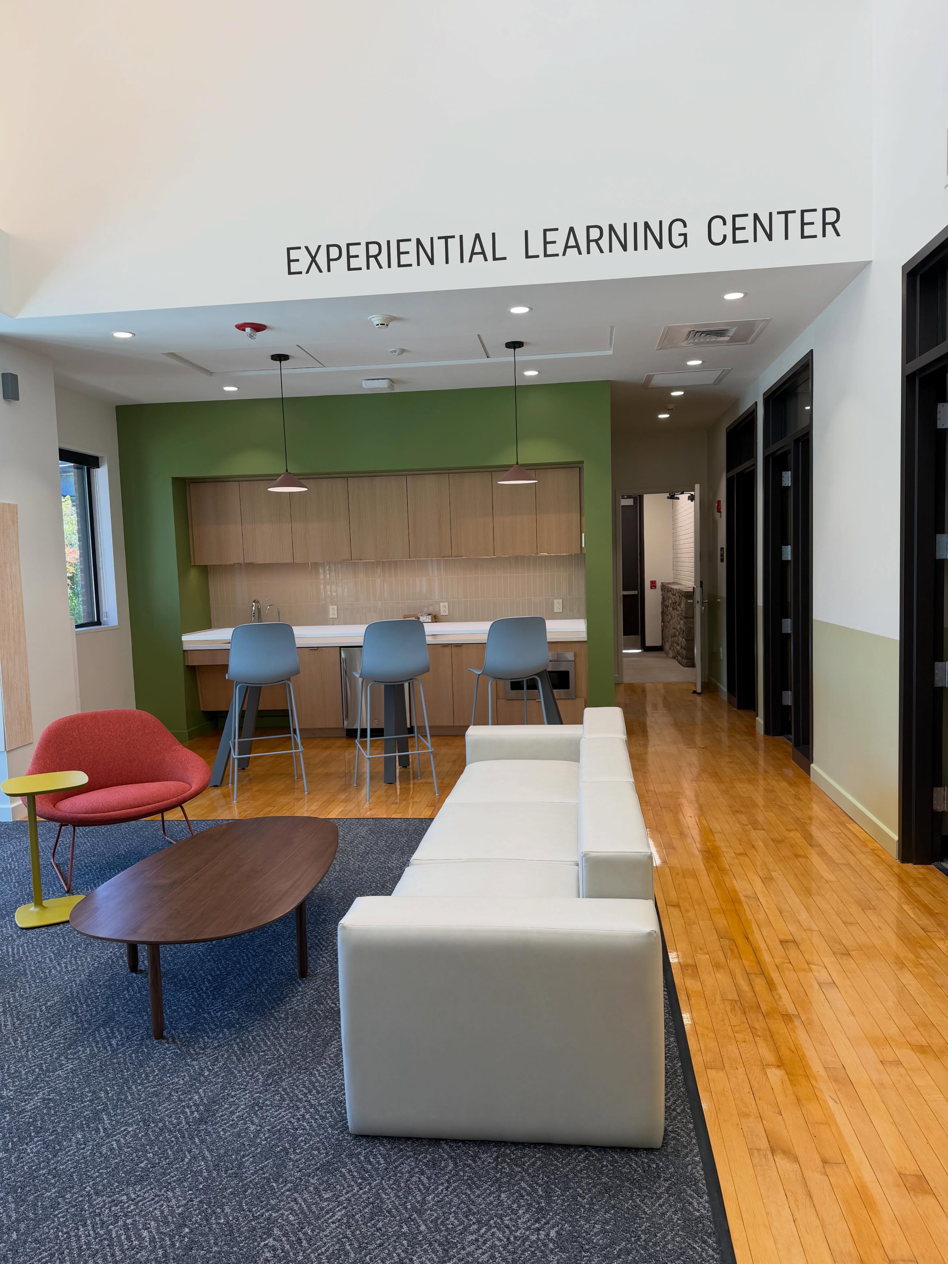 Room labeled "Experiential Learning Center" with a white couch, red chair, and wooden table. A kitchen area with blue stools is in the background.