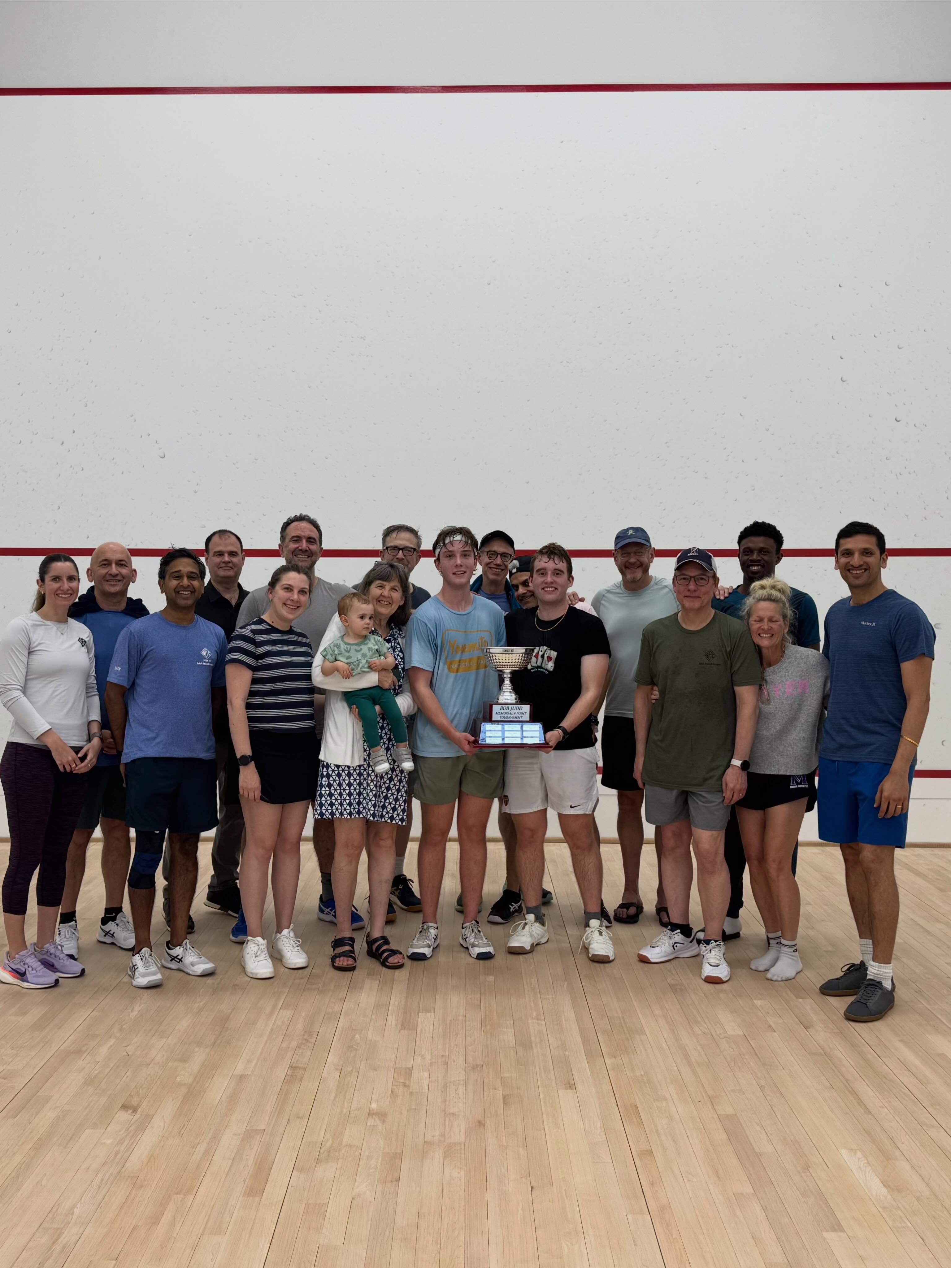 Group of people standing on a squash court, two individuals in the center hold a trophy. Some wear casual clothes and others are in athletic attire.
