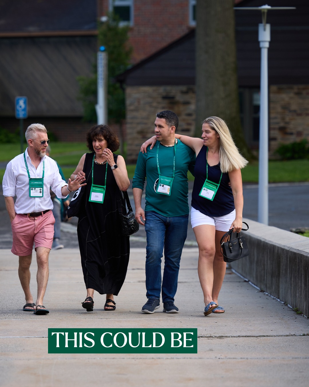 Four people walking on a sidewalk, wearing conference lanyards, engaging in conversation. Brick building and greenery in the background.