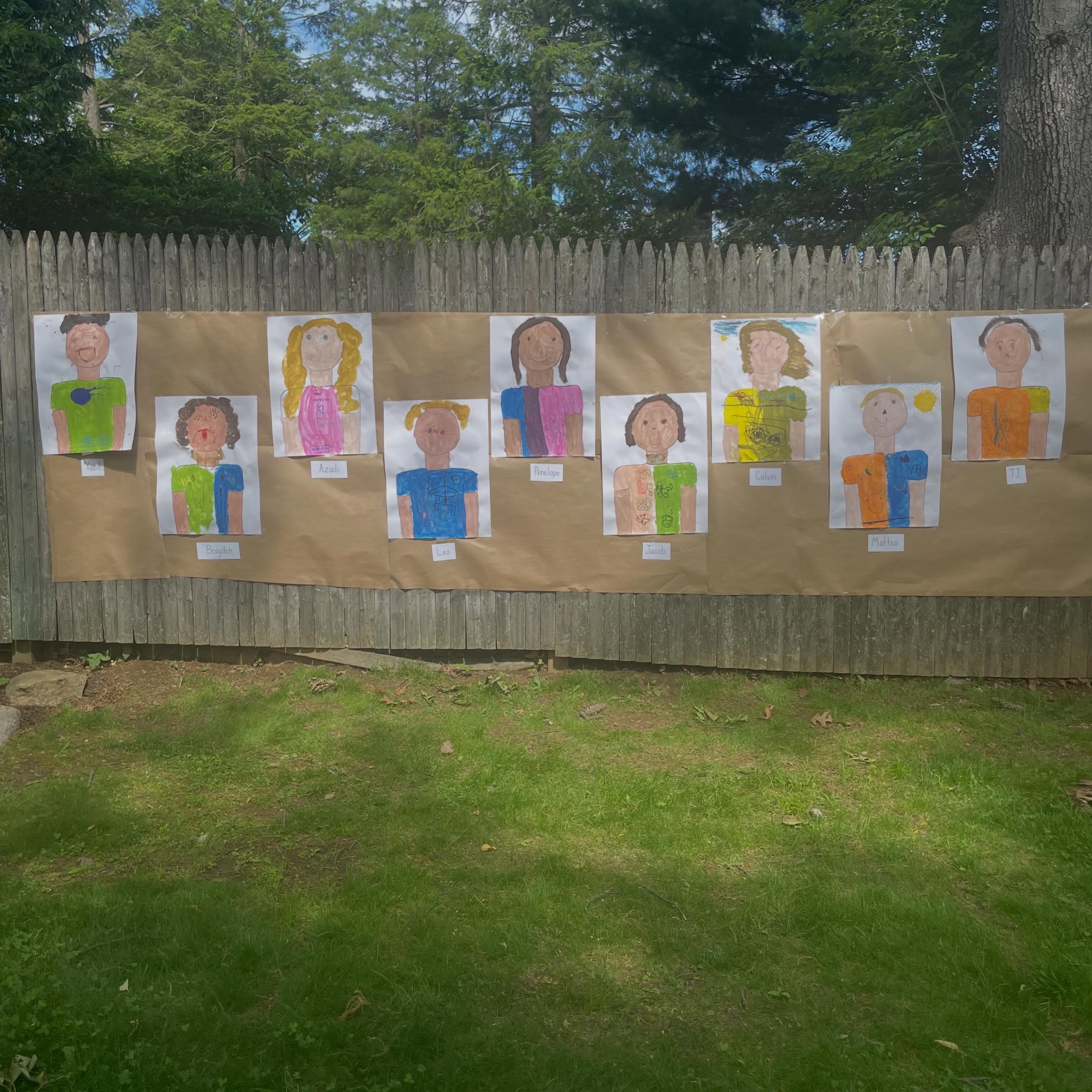 Drawings of people in colorful clothes taped on brown paper against a wooden fence outdoors.