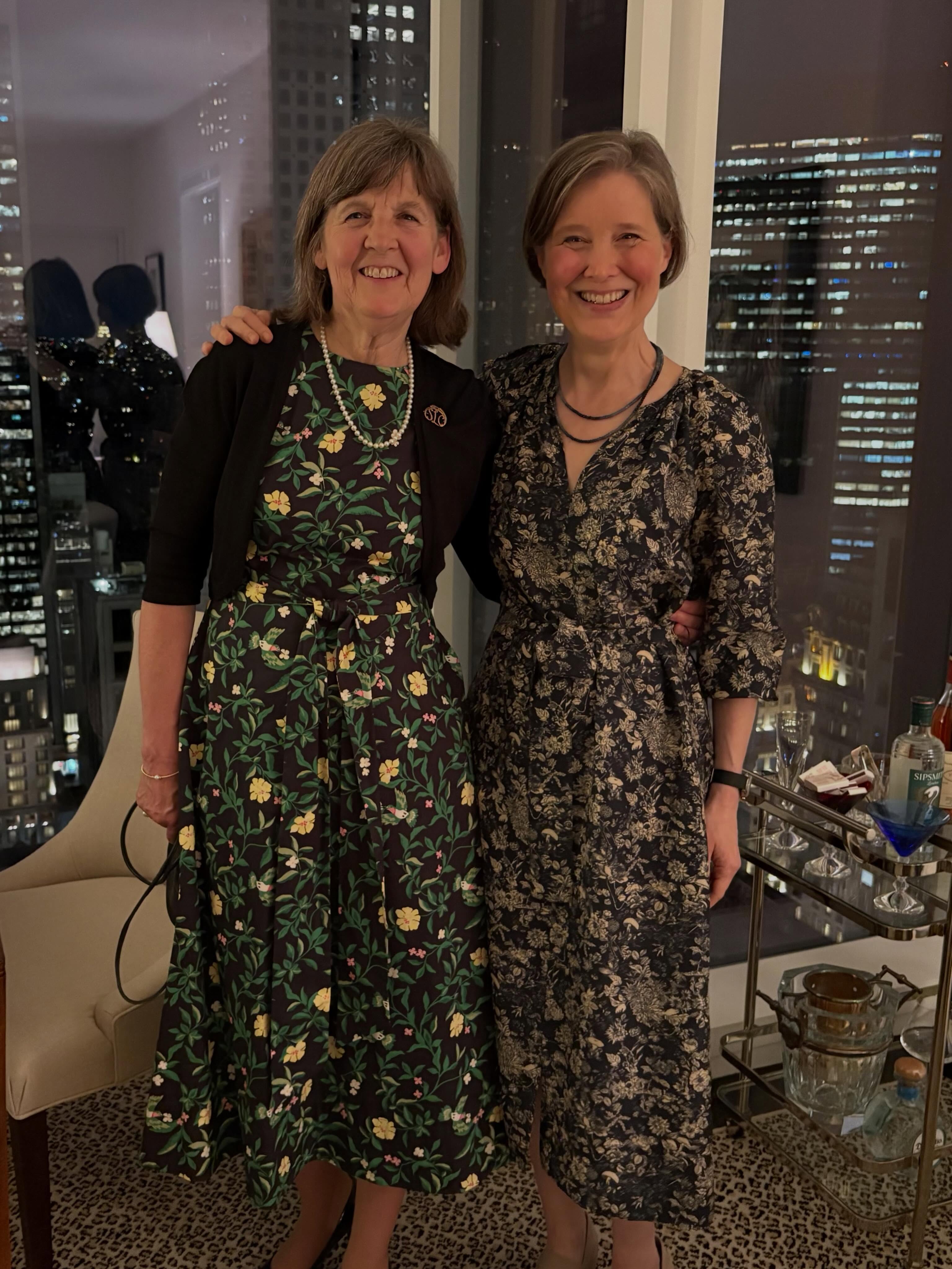 Two people in floral dresses stand smiling in front of a cityscape at night, inside a room with a bar cart and city view.