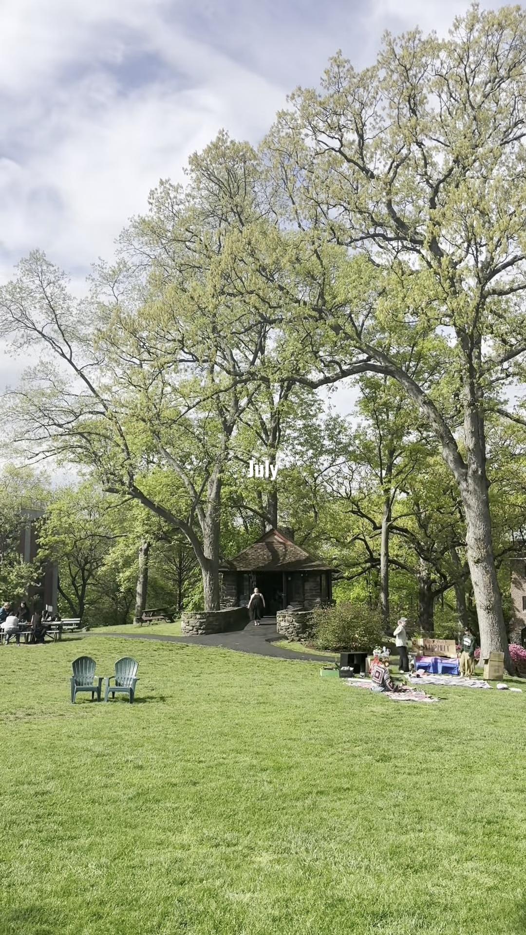 A grassy park with two chairs, large trees, a small stone building, and people near tables. The word "July" is written above.