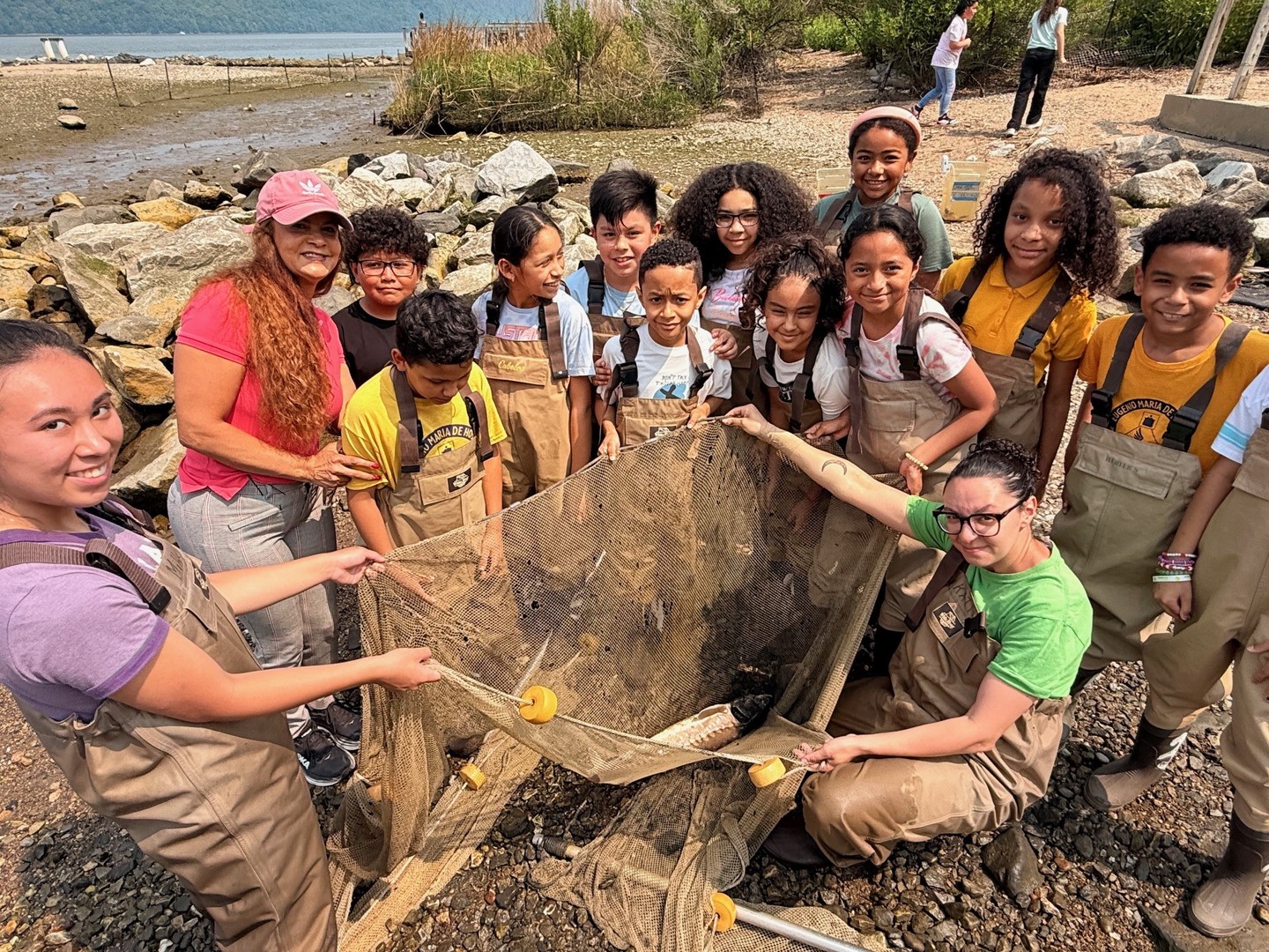 A group of people in waders holding a net by the rocky shore, with water and vegetation in the background.