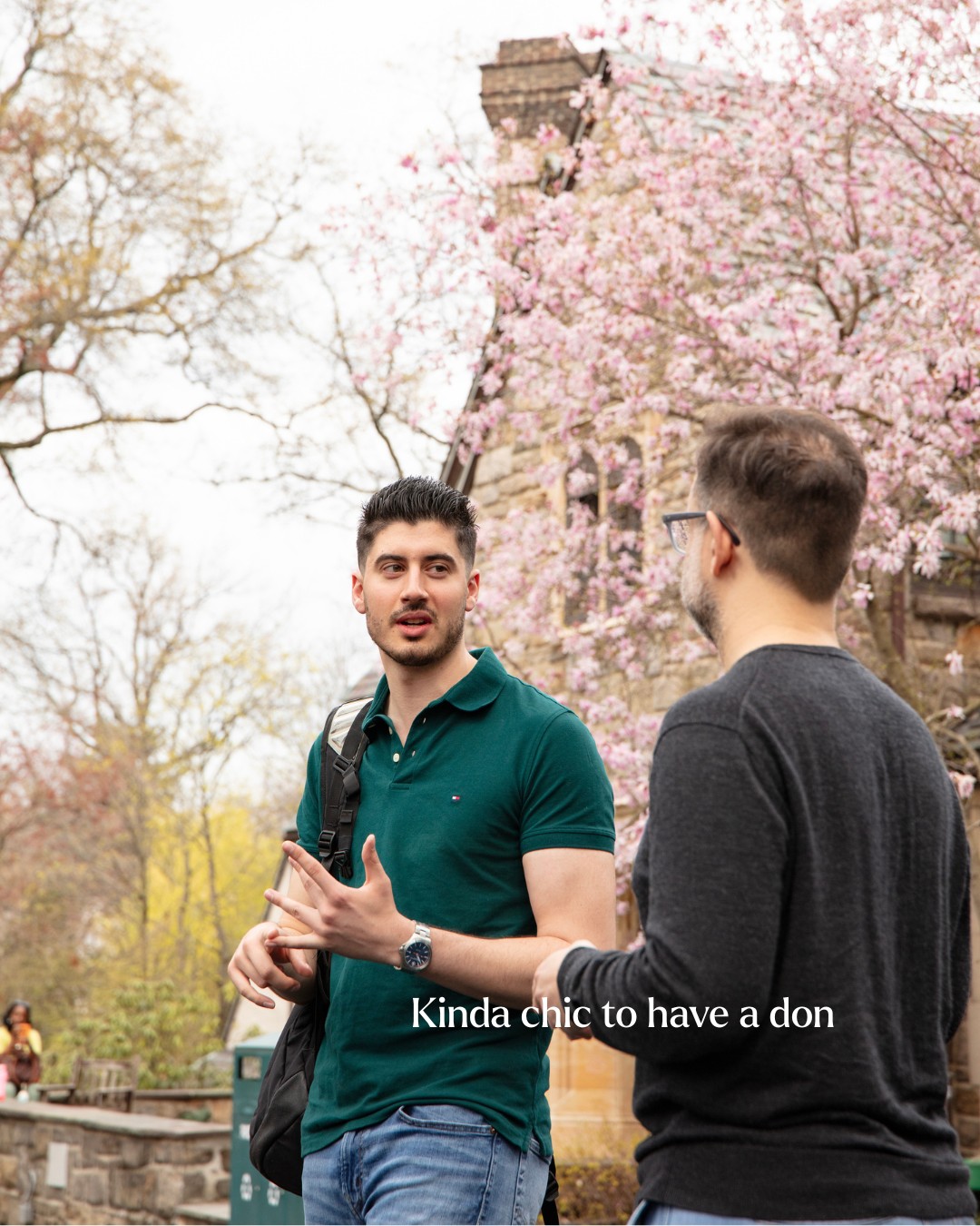 Two people converse outdoors near blossoming trees and an old building. One wears a green shirt, the other a dark sweater. Text reads, "Kinda chic to have a don."