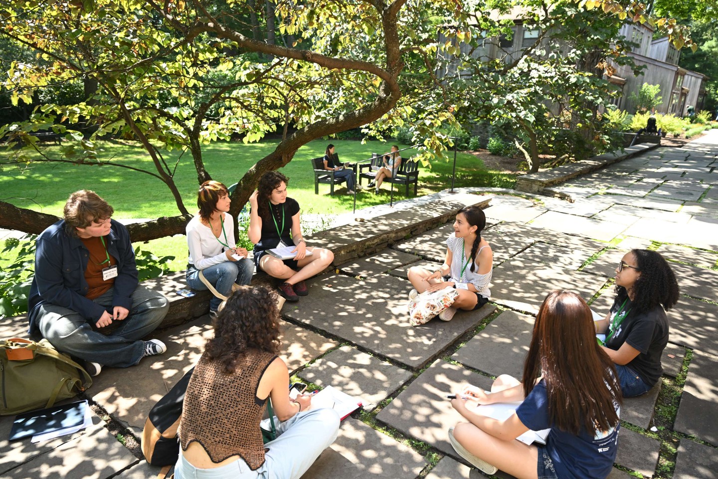 People sitting in a circle on a stone path under a tree, discussing and taking notes, with a garden and more people in the background.