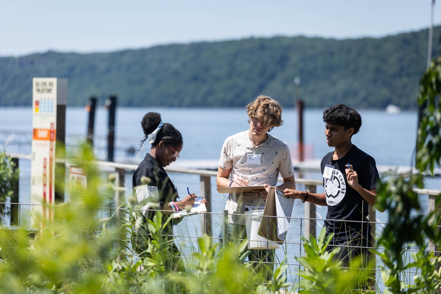 Three people are outdoors by a body of water with trees in the background. They are holding clipboards and writing, surrounded by greenery.