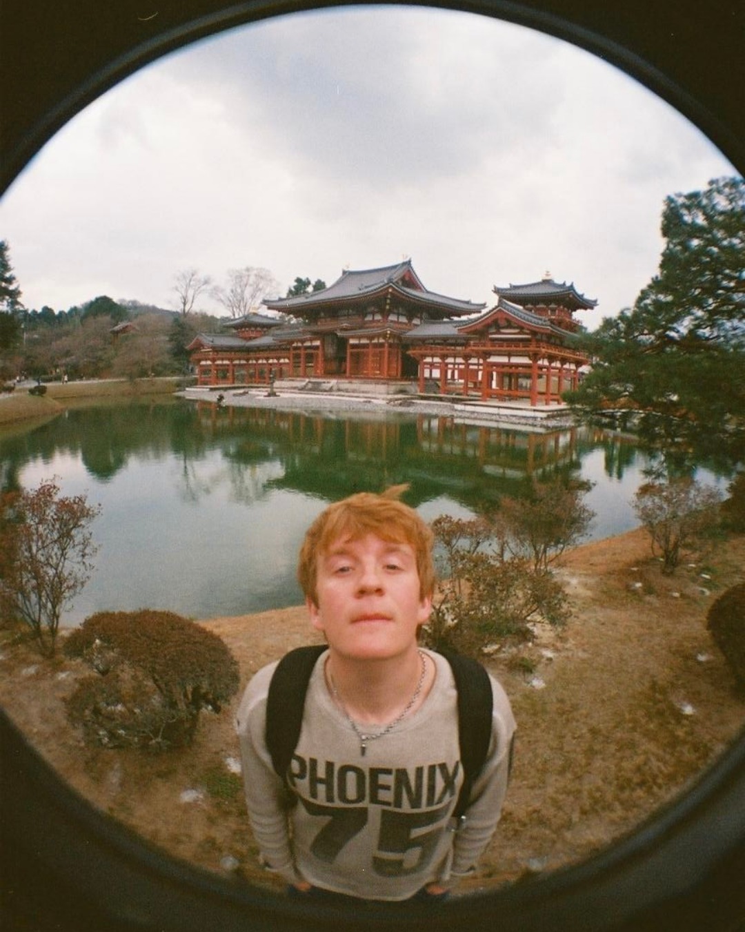 Person wearing a "PHOENIX 75" shirt stands near a serene pond with a traditional Japanese building in the background, viewed through a fisheye lens.