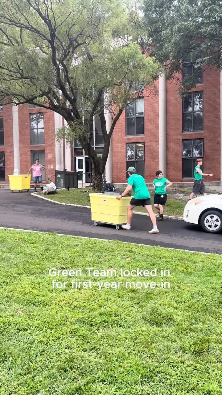 People in green shirts moving large yellow carts on a path by a brick building, text reads "Green Team locked in for first-year move-in."