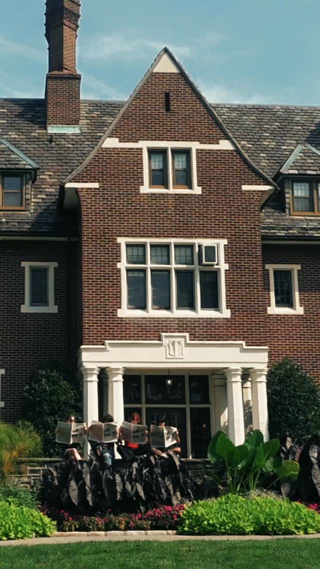 A group of seated people read newspapers in front of a large brick house with decorative windows and a lush garden.