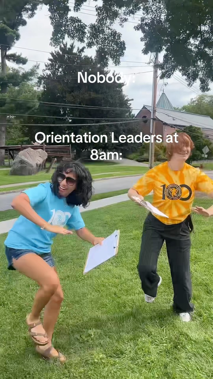 Two people on grass, one in a blue shirt and shorts holding a clipboard, the other in a yellow shirt and black pants, both dancing energetically.