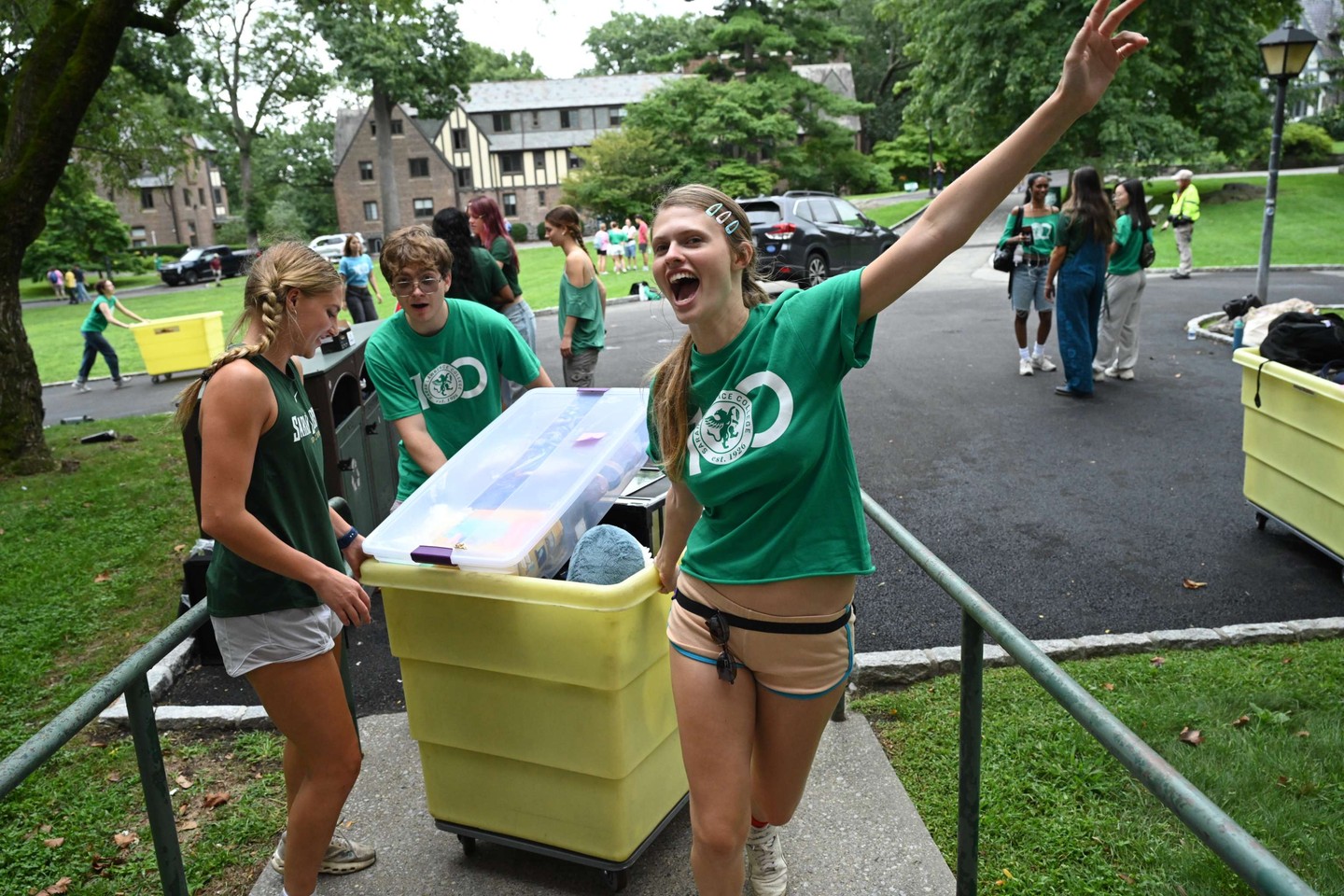 Students in green shirts move yellow bins on a campus. One smiles and raises an arm, surrounded by trees and buildings in the background.