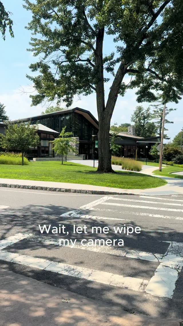 Modern building surrounded by trees and a crosswalk in the foreground. Text on image reads, "Wait, let me wipe my camera."