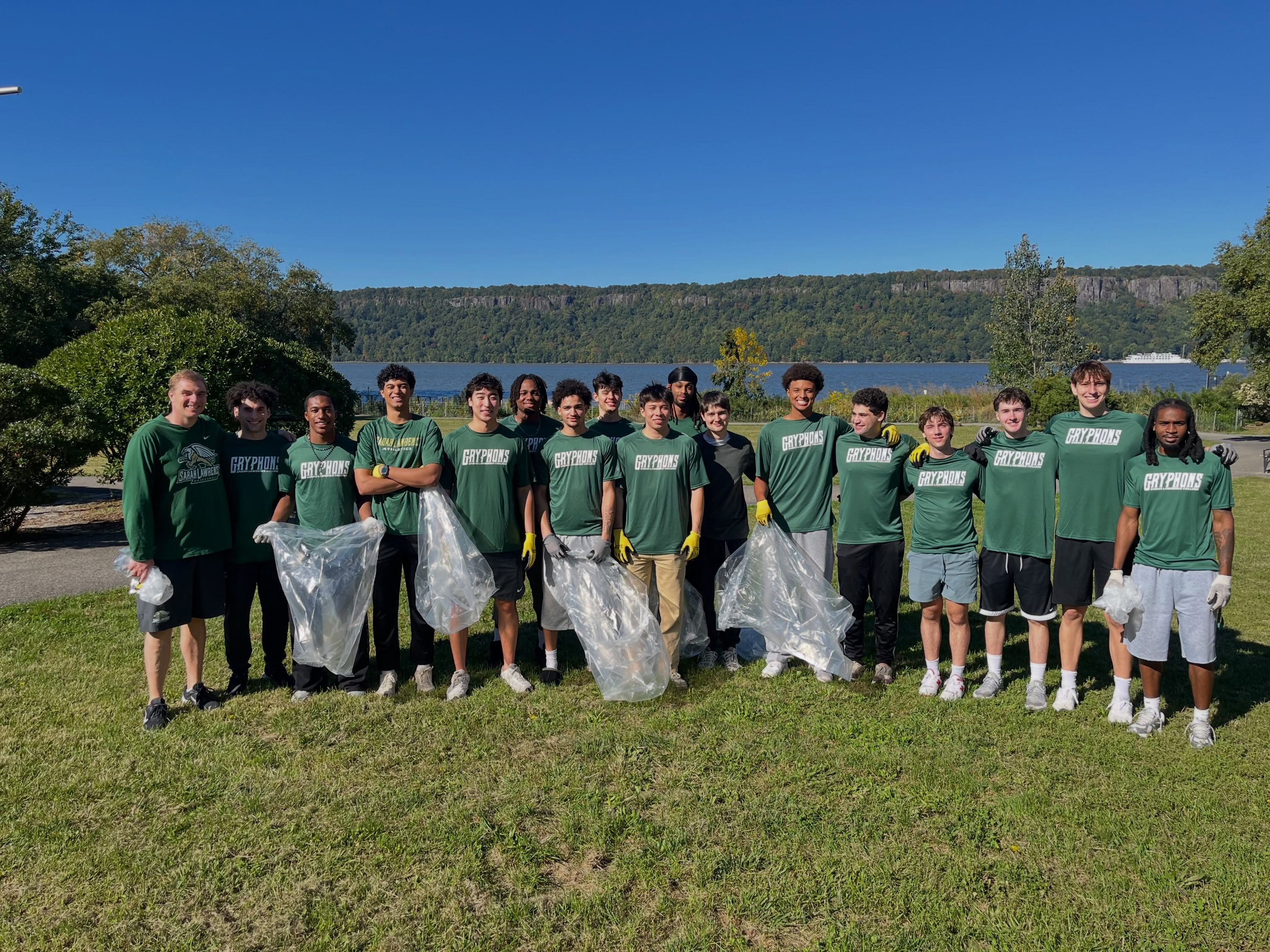 A group wearing matching green shirts poses outdoors with clear trash bags, in front of a scenic view of water and trees under a clear blue sky.