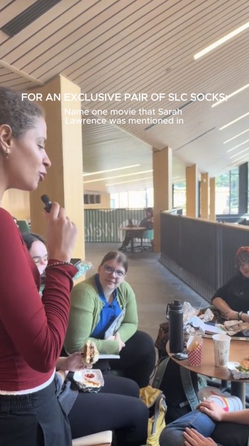 Person speaking with a microphone to a group seated with food. Text on image reads: "FOR AN EXCLUSIVE PAIR OF SLC SOCKS."