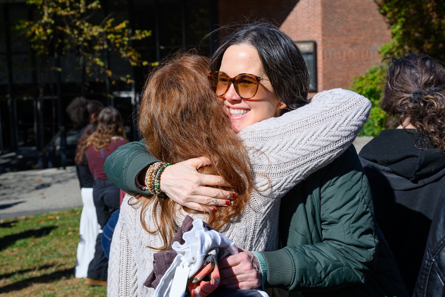 Two people warmly hug outdoors on a sunny day. One wears sunglasses and a green jacket; the other has long hair and a knitted sweater.