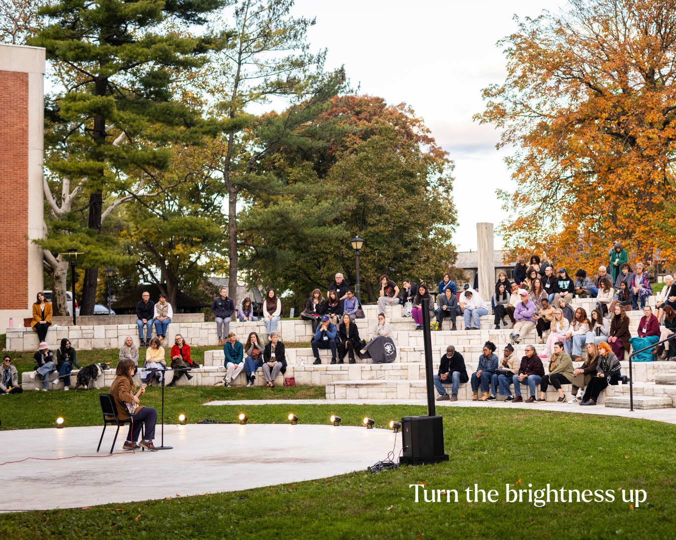 Outdoor performance with a seated performer in front of an amphitheater audience. Trees and autumn foliage in the background.