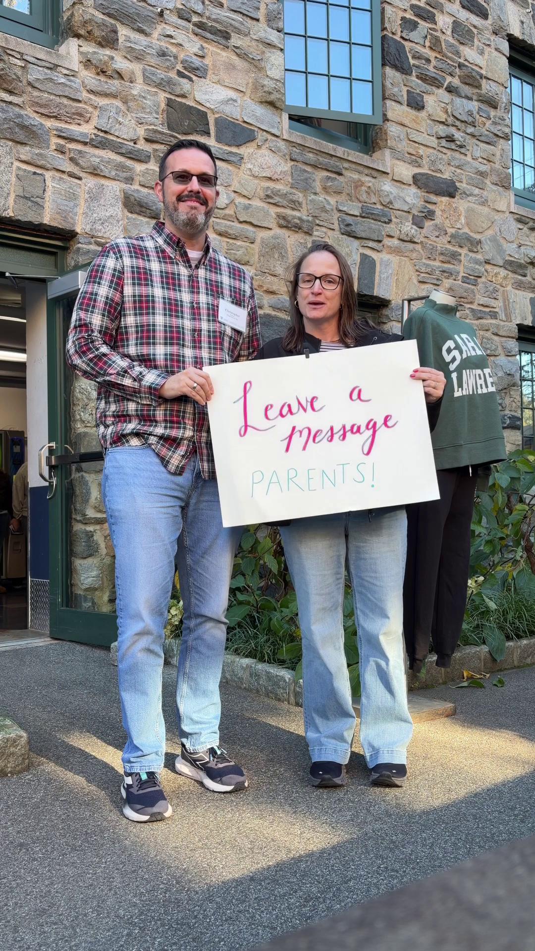 Two people stand holding a sign that says, "Leave a message parents!" in front of a stone building.