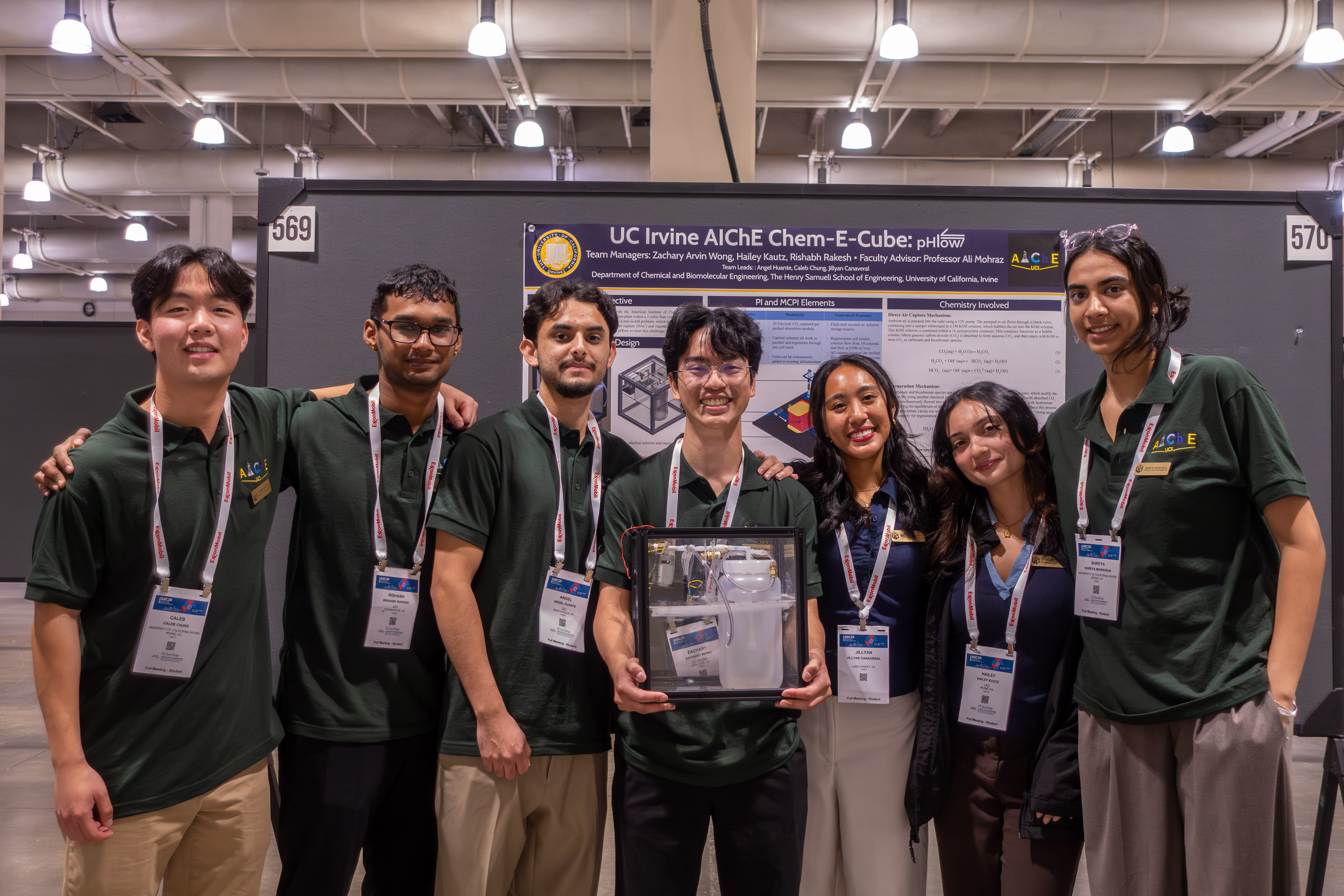 Seven members of the ChemE Cube team pose with their cube, wearing matching green T-shirts. Behind them is a detailed information board for their presentation.