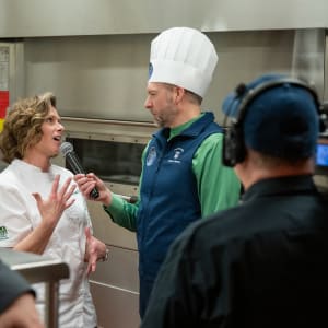 Chef Puff in a white coat speaks passionately into a microphone during an interview at the 2024 Celebrity Chefs Invitational. The interviewer, wearing a chef’s hat and vest, listens attentively, while a cameraman captures the moment in a professional kitchen setting.