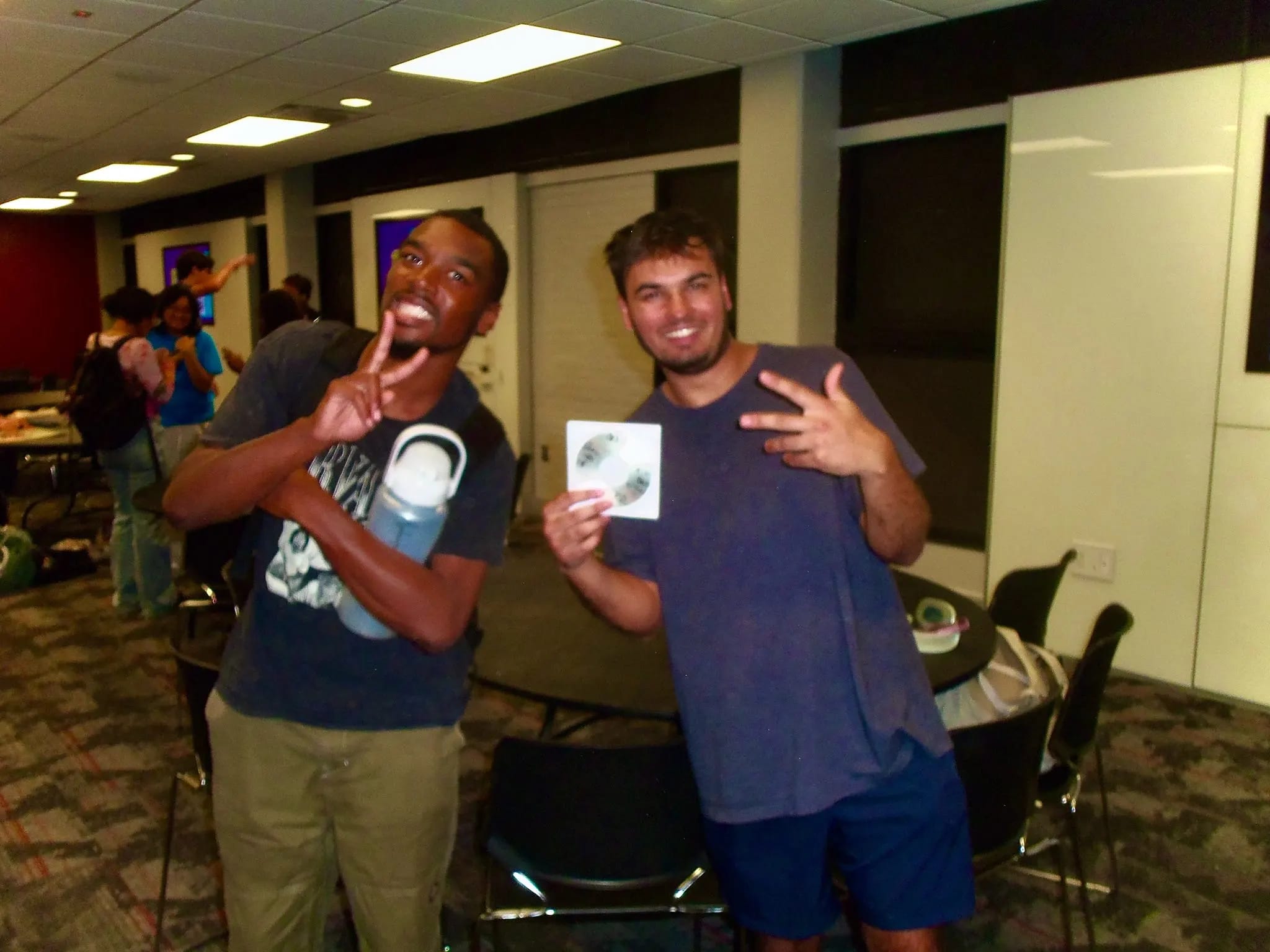 Two people smiling and posing indoors; one holds a CD while both make peace signs near tables and chairs.