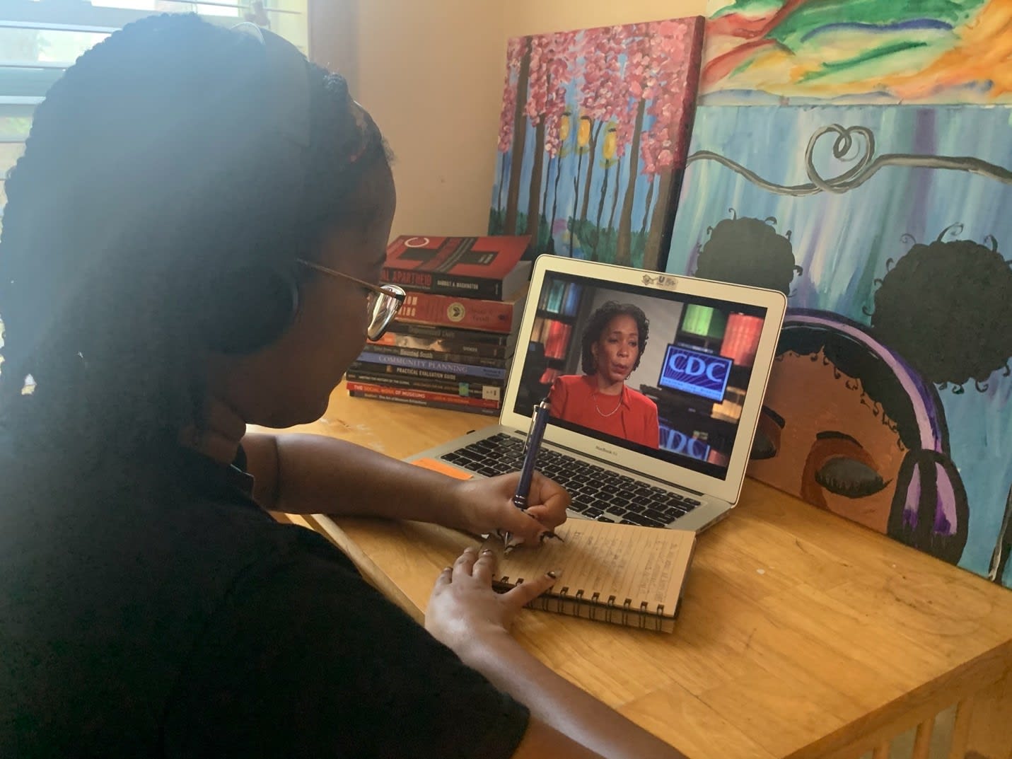 Deniseya Hall sits at a desk with art behind the computer, taking notes on a notebook while watching a presentation on a laptop from the CDC.