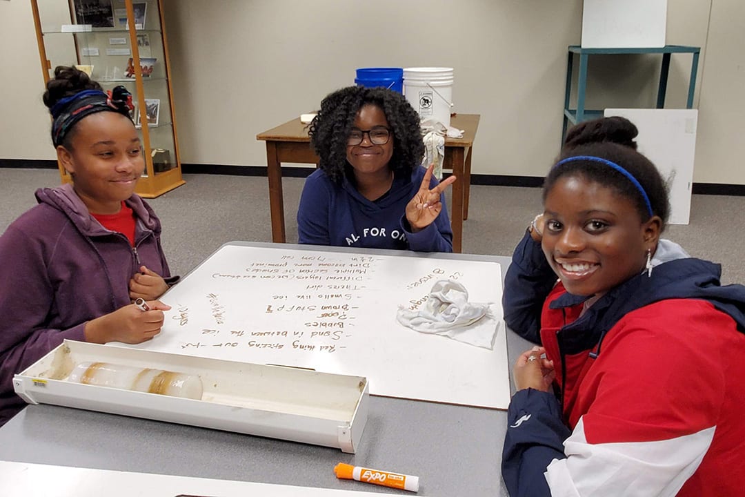 Three young students sit together at a table, working on a poster 