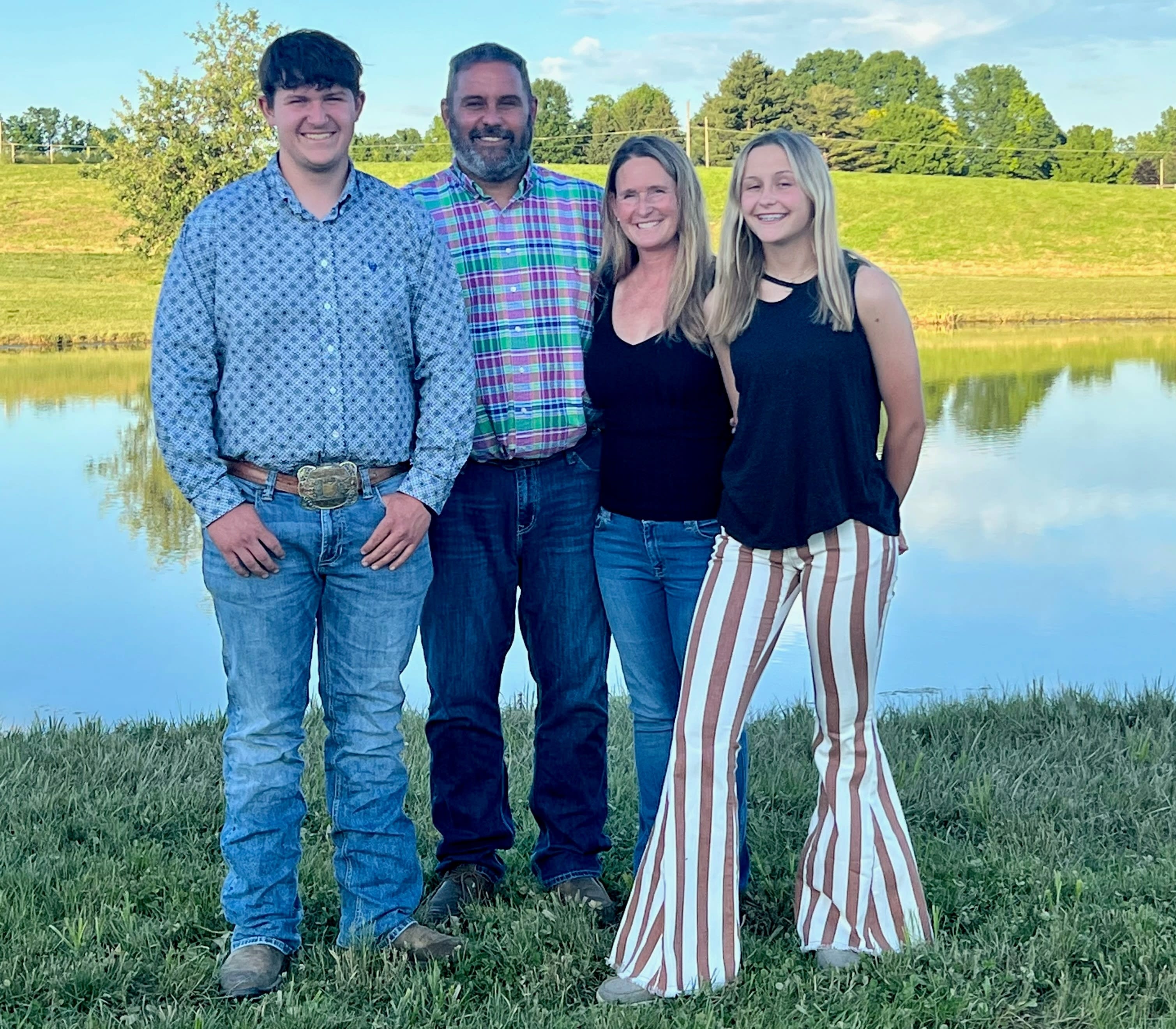 Four members of the Love family standing outside. They are posed at the edge of a lake with a large green patch behind it