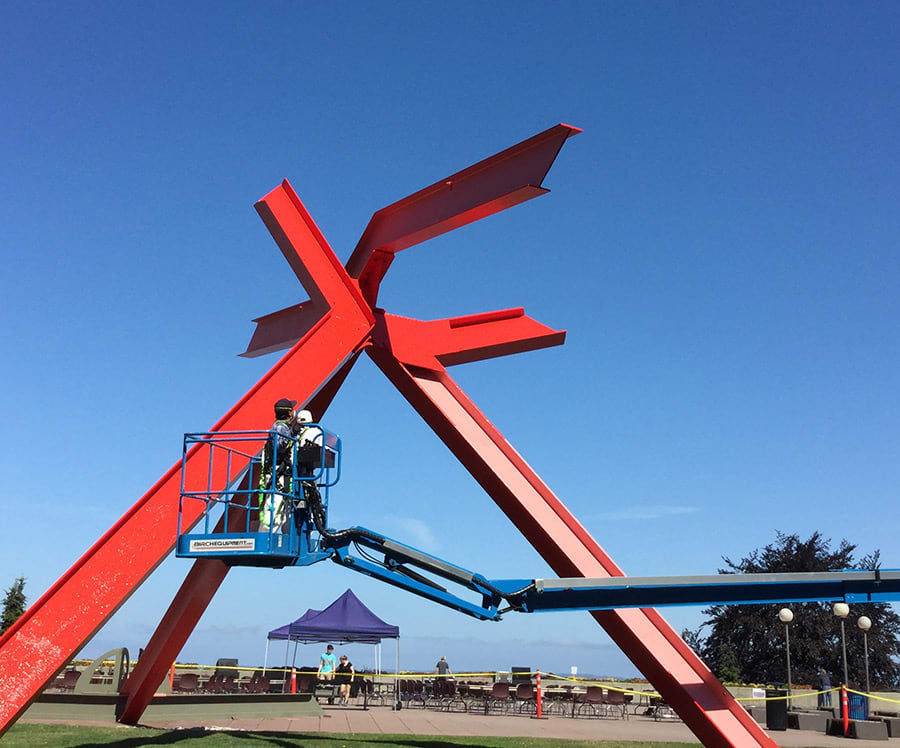 large red metal sculpture being painted by two people in a lift