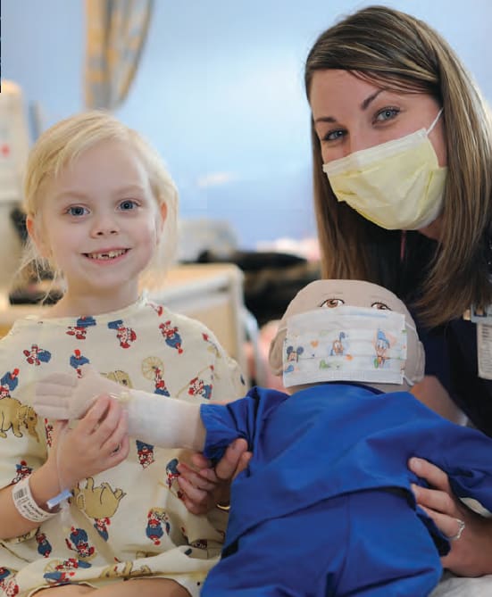 A young girl and a nurse smile as they both hold a doll wearing a surgical mask,