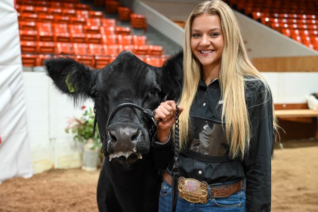 A girl stands in an arena, holding the reigns of her black cow. She smiles towards the camera